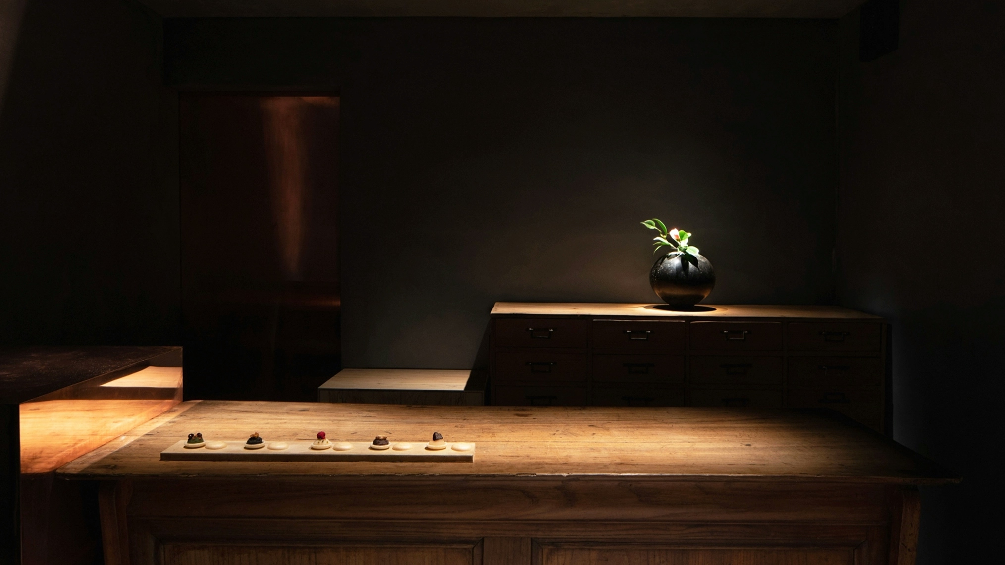 A dark room with a low wooden counter in the foreground has a long, thin plate with five sweets delicately placed atop. In the background a dark vase sits in the shadows with a refined floral arrangement